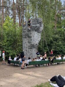 Grupa uczniów siedzi na ławkach. W środku skałka wspinaczkowa, na którą wspinaj się jedna osoba. 
