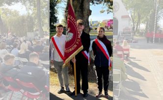Troje uczniów na tle osób siedzących na uroczystości. Uczniowie mają na sobie szarfy zw kolorze biało-czerwonym, środkowy uczeń trzyma sztandar szkoły.