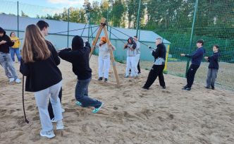 Grupa uczniów na piaszczystym boisku uczestniczy w grze terenowej.