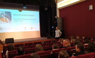 Sala teatralna, na czerwonych krzesełkach siedzi kilka osób. Z przodu wykładowca i ekran z prezentacją.