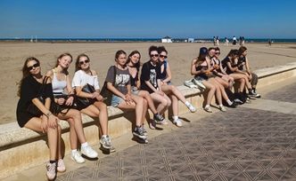 Kilkanaście osób siedzi na murku na deptaku przy plaży.