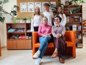 Na pomarańczowej sofie siedzą dwie nauczycielki, z tyłu za sofą stoi troje uczniów. Tło to regały biblioteczne i gazetka. 