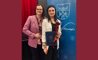 Dwie kobiety na tle ściamki promocyjnej Powiatu Wołomińskiego. Jedna trzyma różę i teczkę.