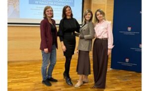 Cztery kobiety stoją na środku sali konferencyjnej. W tle ekran z tytułem konferencji.