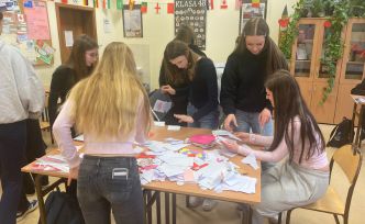 Grupa młodych osób pochylonych nad stołem w szkolnej sali, sortujących listy walentynkowe.