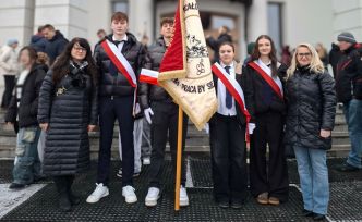 Grupa młodych osób i dwie dorosłe osoby stoją razem przed budynkiem kościoła, trzymając szkolny sztandar.