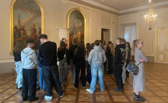 Grupa młodych osób stojąca w muzeum w zabytkowej sali z dużymi obrazami na ścianach.