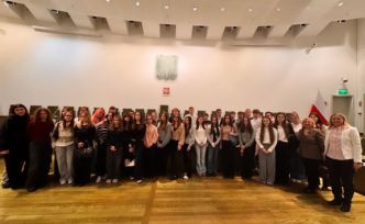 Grupa osób stoi razem w dużej, jasnej sali. Na ścianie wisi godło i flaga.