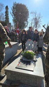 Młodzież i nauczyciele oddają hołd podczas uroczystości na cmentarzu, w tle sztandar szkoły.