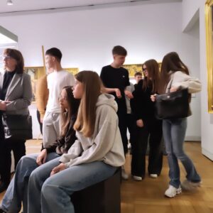 Grupa młodzieży (część stoi , część siedzi na pufach) słucha wykładu w muzeum. 