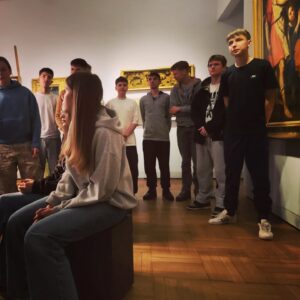 Grupa młodzieży (część stoi , część siedzi na pufach) słucha wykładu w muzeum. 