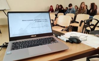 Laptop na biurku z prezentacją o prawie autorskim, a w tle grupa uczniów czekających na zajęcia.