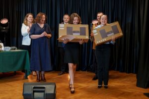 Dwie uczestniczki konkursu trzymają w rękach kartony z nagrodami. W tle organizatorzy . 