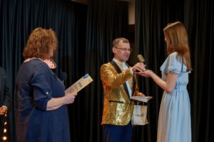 Mężczyzna w złotej marynarce wręcza nagrodę (statuetka) uczestniczce konkursu. Kobieta obok wręcza dyplom.