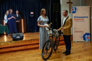 Mężczyzna w złotej marynarce wręcza nagrodę (rower) uczestniczce konkursu.