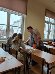 Uczniowie siedzą w klasie lekcyjnej, przy nich koło ławki prowadzący zajęcia