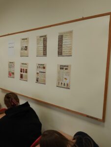 Gazetka szkolna z widocznymi plakatami. W rogu uczeń siedzący przy ławce szkolnej.