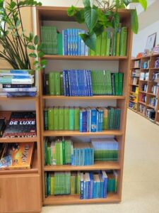 Regał z książkami zapełniony książkami zielonym i niebieskimi. Niebieskie układają się k kształt rzeki. 