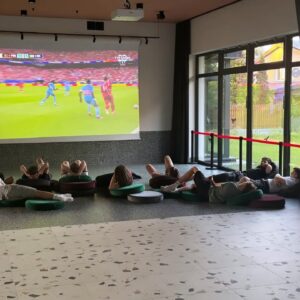 Kilka osób na dużej sali ogląda mecz na ekranie.