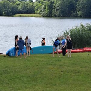 Na pierwszym planie trawiasta plaża przy jeziorze. Nad jeziorem kilka osób z kajakami.
